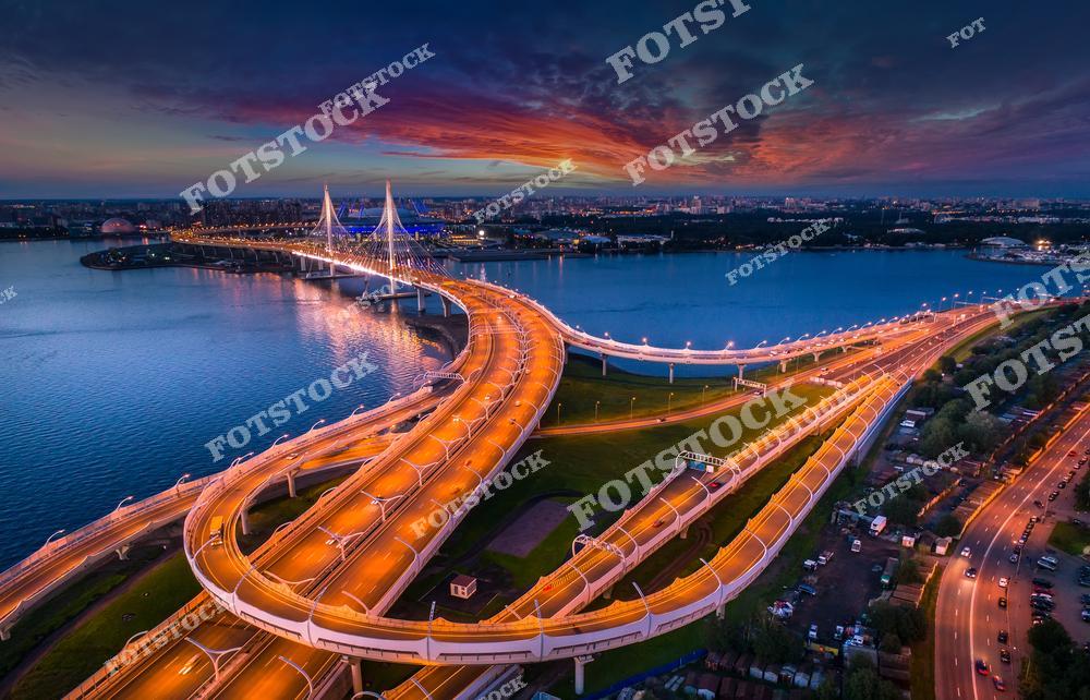 На снимке изображен Западный скоростной диаметр, пересекающий Финский залив. Вантовый мост и изящные линии развязок подсвечены вечерними огнями. Закатное небо с насыщенными красками отражается в водах залива, добавляя снимку драматичности. Этот вид символизирует современную транспортную архитектуру Санкт-Петербурга.